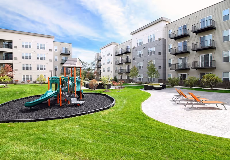 Outdoor courtyard area of a senior living facility with green grass, a children's playground with a slide and climbing structure, several lounge chairs on a paved patio, and multi-story residential buildings with balconies in the background under a partly cloudy sky.