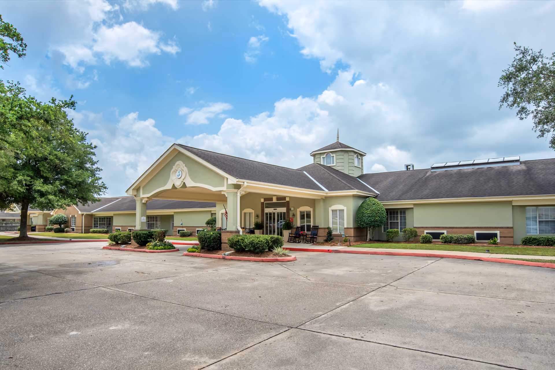 Front entrance of a single-story senior living facility with a covered porte-cochere, landscaped beds, and a driveway under a partly cloudy sky.