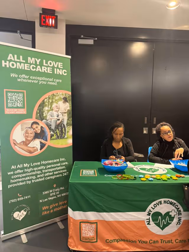 Two staff sit at a promotional booth with a green All My Love Home Care banner.