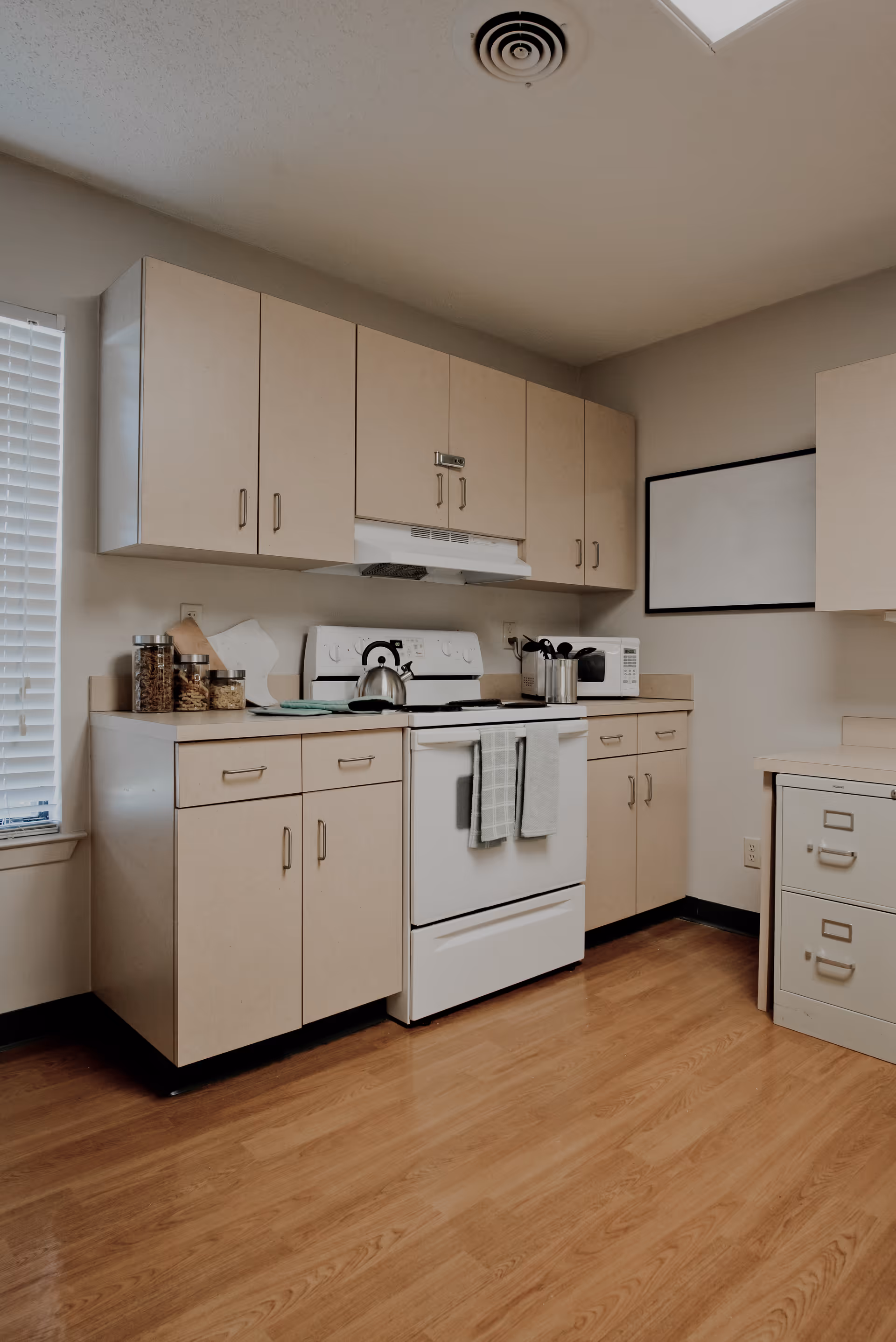 A clean kitchen area with beige cabinets, a white stove with a kettle on it, a microwave, and various kitchen utensils on the countertop. The floor is wooden, and there is a window with blinds on the left side.