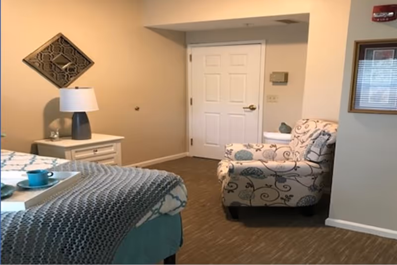Bedroom in senior living facility with bed, nightstand, lamp, patterned chair, and doorway.