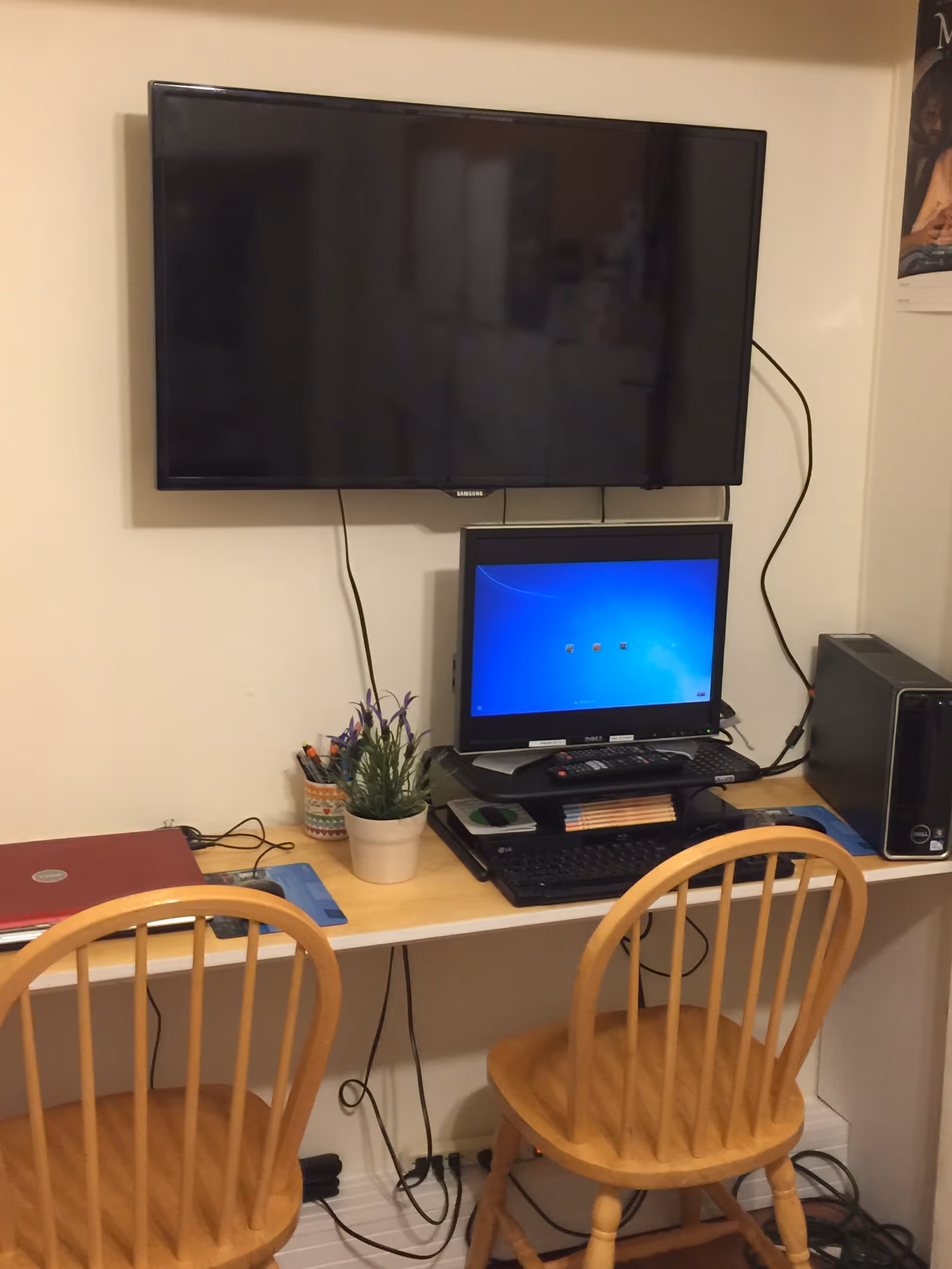 Desk with wall-mounted TV, monitor, laptop, plant, and two wooden chairs.