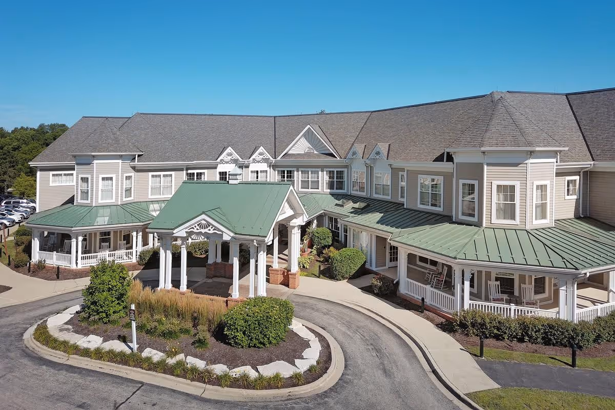 Front exterior of a two-story senior living facility with green metal roofs, a covered portico entrance, wraparound porches and a circular driveway.