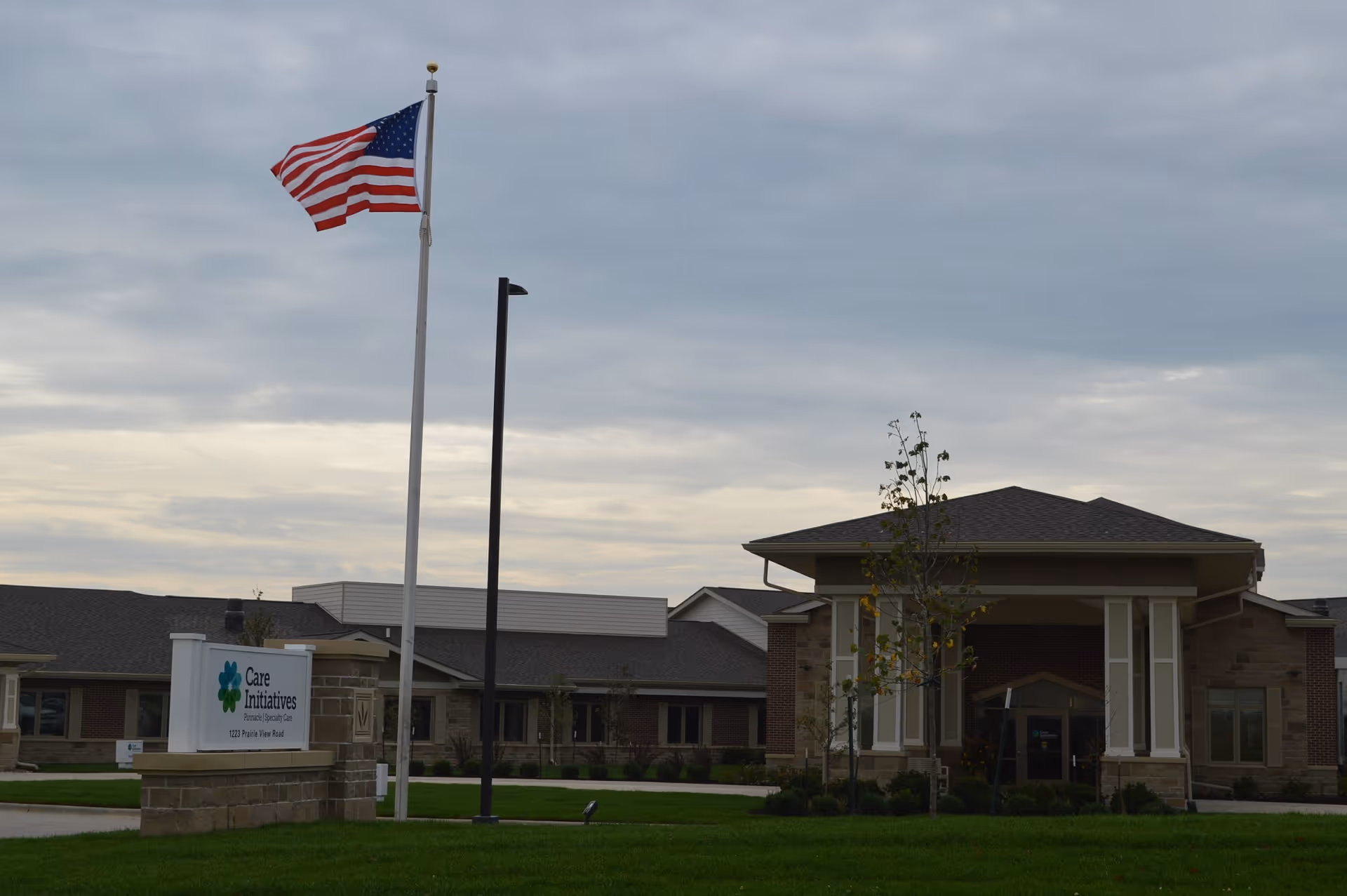 Exterior view of a senior living facility with a large American flag on a flagpole in front. The building has a covered entrance with columns and a well-maintained lawn. A sign near the entrance reads 'Care Initiatives'. The sky is overcast.
