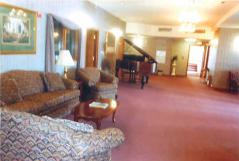 A spacious common area with patterned upholstered sofas and armchairs arranged around a wooden coffee table. A grand piano is visible in the background along the far wall. The room has carpeted flooring, wall-mounted lights, framed artwork, and a window with curtains.