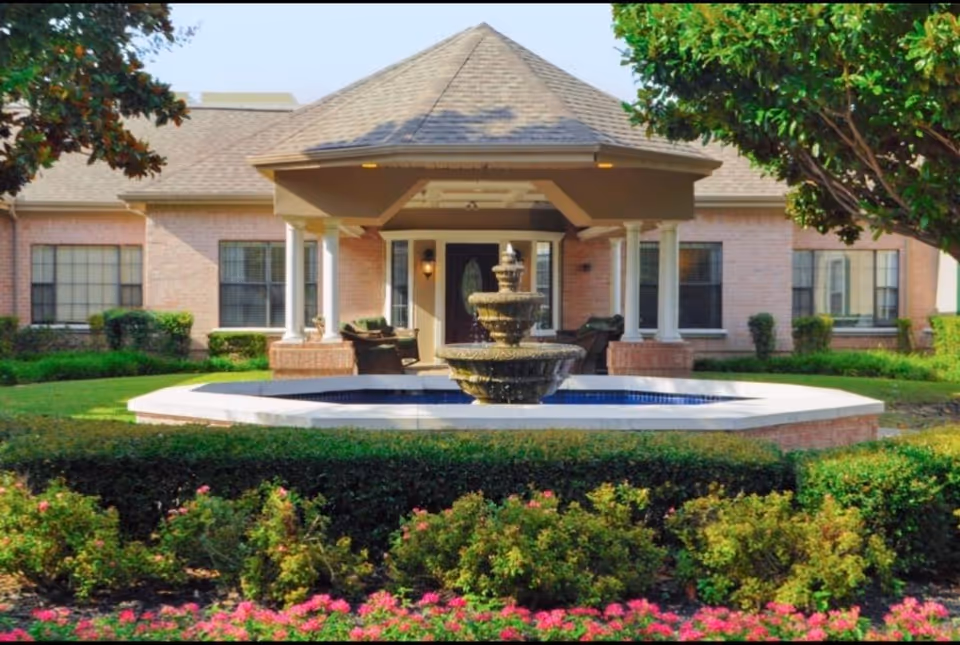 Front entrance of a brick senior living building with a covered portico, central tiered fountain, and manicured landscaping.