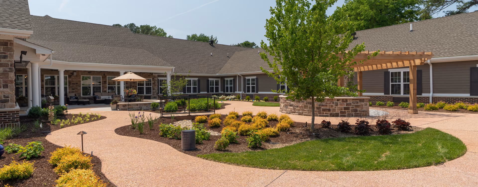 Outdoor courtyard area at a senior living facility with a paved walking path, landscaped garden beds with shrubs and small trees, a wooden pergola, and a building with multiple windows and a covered patio with seating and an umbrella.