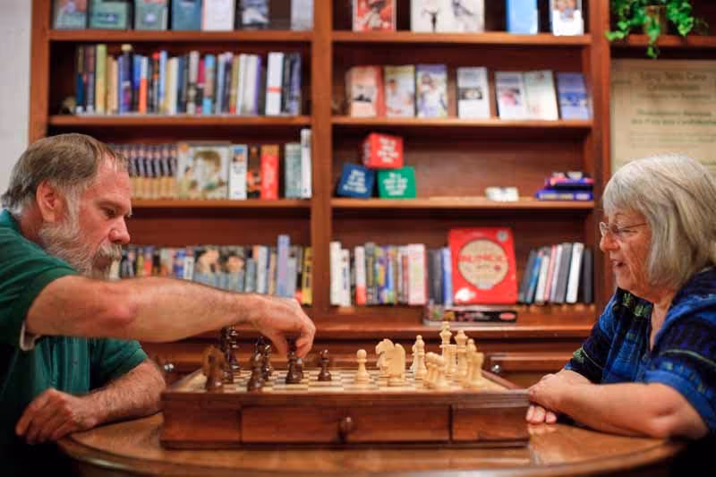 An elderly man and woman playing chess at a wooden table in a cozy room with bookshelves filled with books and games in the background.