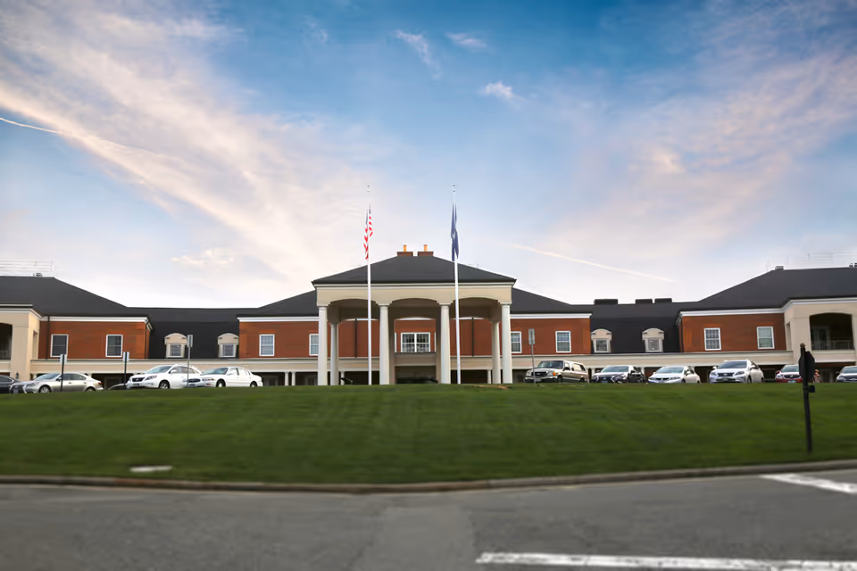 Front exterior of a senior living building with a columned portico, flagpoles, a grassy lawn, and parked cars.