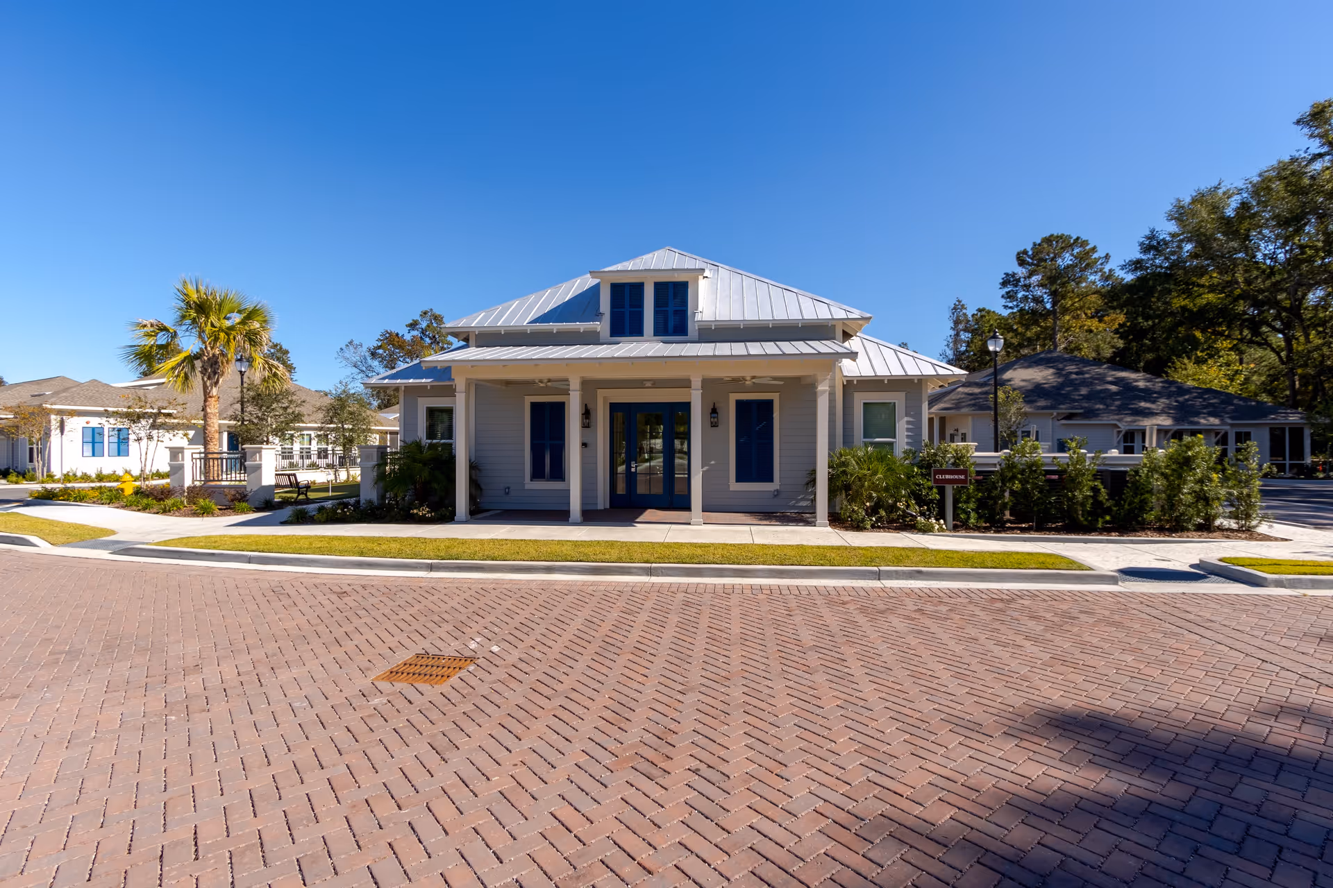 Front exterior view of a single-story building with a metal roof, blue doors and windows, surrounded by landscaping including palm trees and bushes, under a clear blue sky.