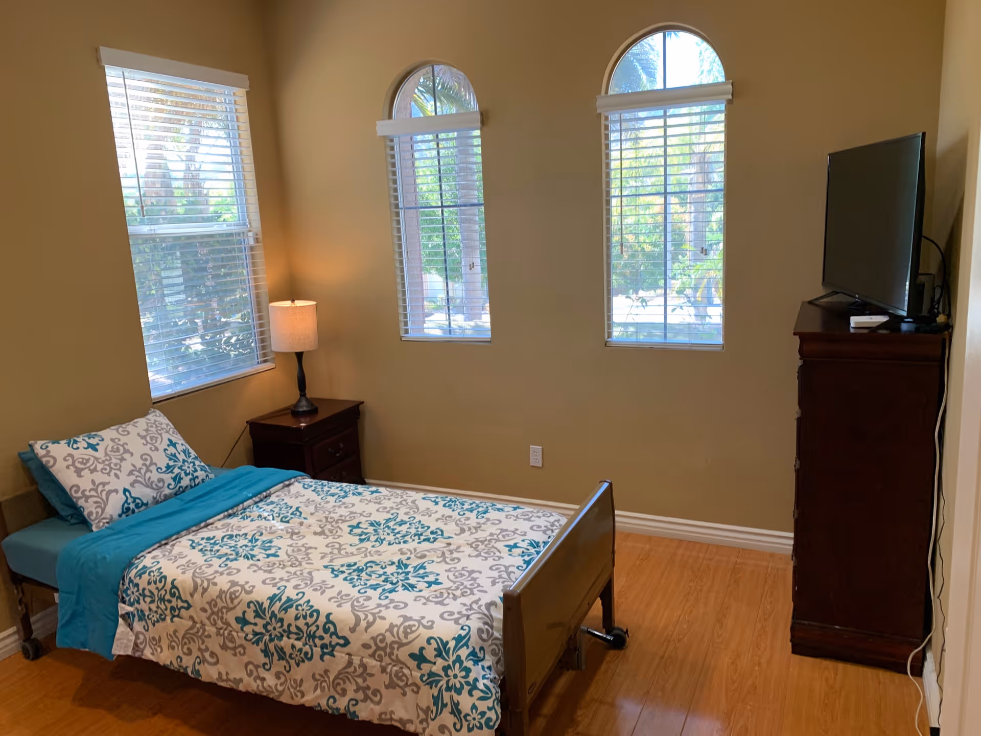 A bedroom with a single bed covered in a white and teal patterned bedspread and matching pillow. Next to the bed is a dark wooden nightstand with a lamp. The room has three windows with blinds, two of which are arched. A dark wooden dresser with a flat-screen TV on top is positioned against the wall. The floor is wooden, and the walls are painted beige.