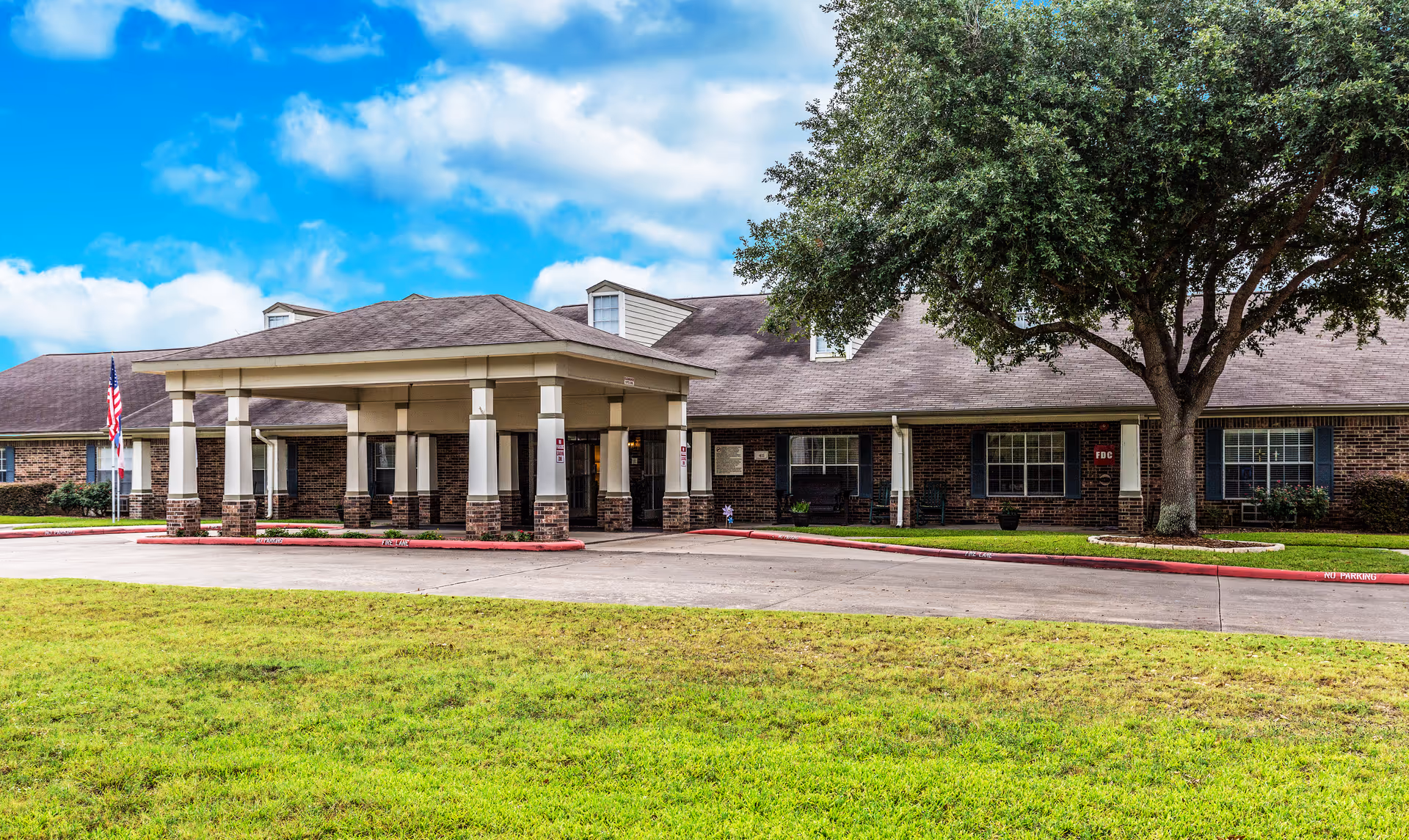 Front entrance of a single-story brick assisted living facility with a covered porte-cochère, lawn, and a large tree.