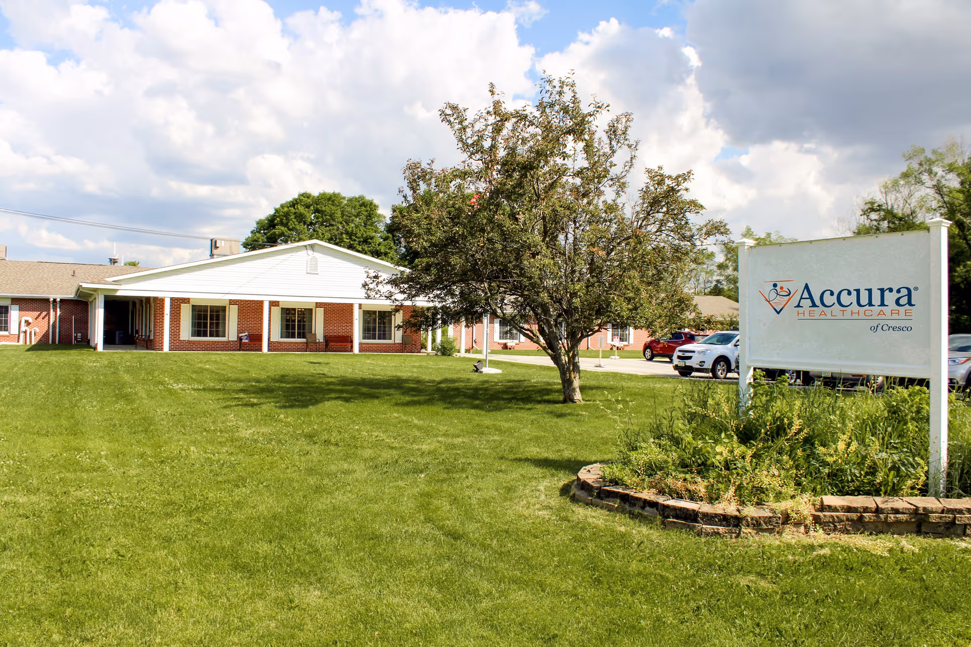 Front lawn and entrance of Accura HealthCare of Cresco showing the building facade, a tree, parked cars, and a large sign.