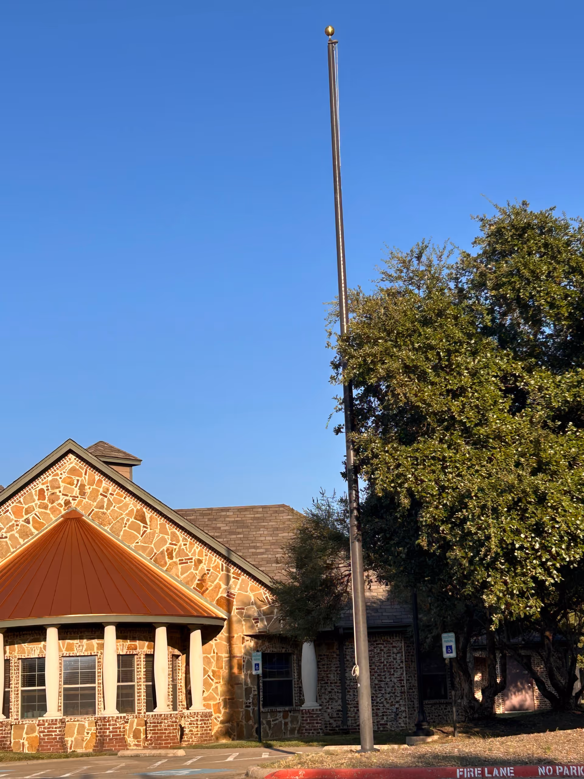 Stone and brick facility facade with a tall flagpole, orange roofed entrance, trees, blue sky.