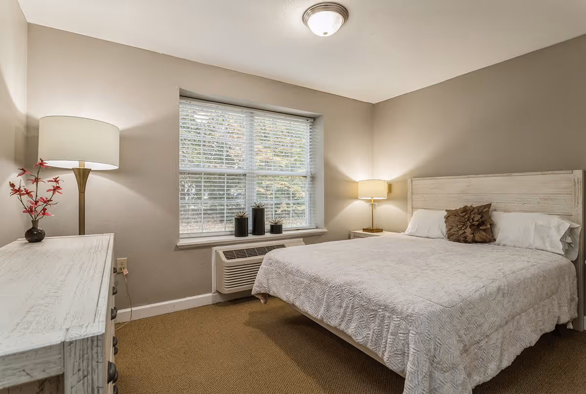 A cozy bedroom with a large bed covered in a white textured bedspread and a decorative brown pillow. There is a white wooden headboard, a nightstand with a lamp on the right side of the bed, and a dresser with a lamp and a small vase with red flowers on the left side. A window with white blinds lets in natural light, and an air conditioning unit is installed below the window. The walls are painted beige and the carpet is light brown.