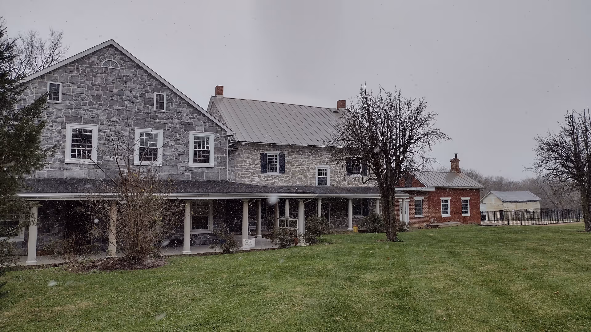 Stone Evergreen Homestead exterior with a long porch, lawn, and leafless trees in snow.