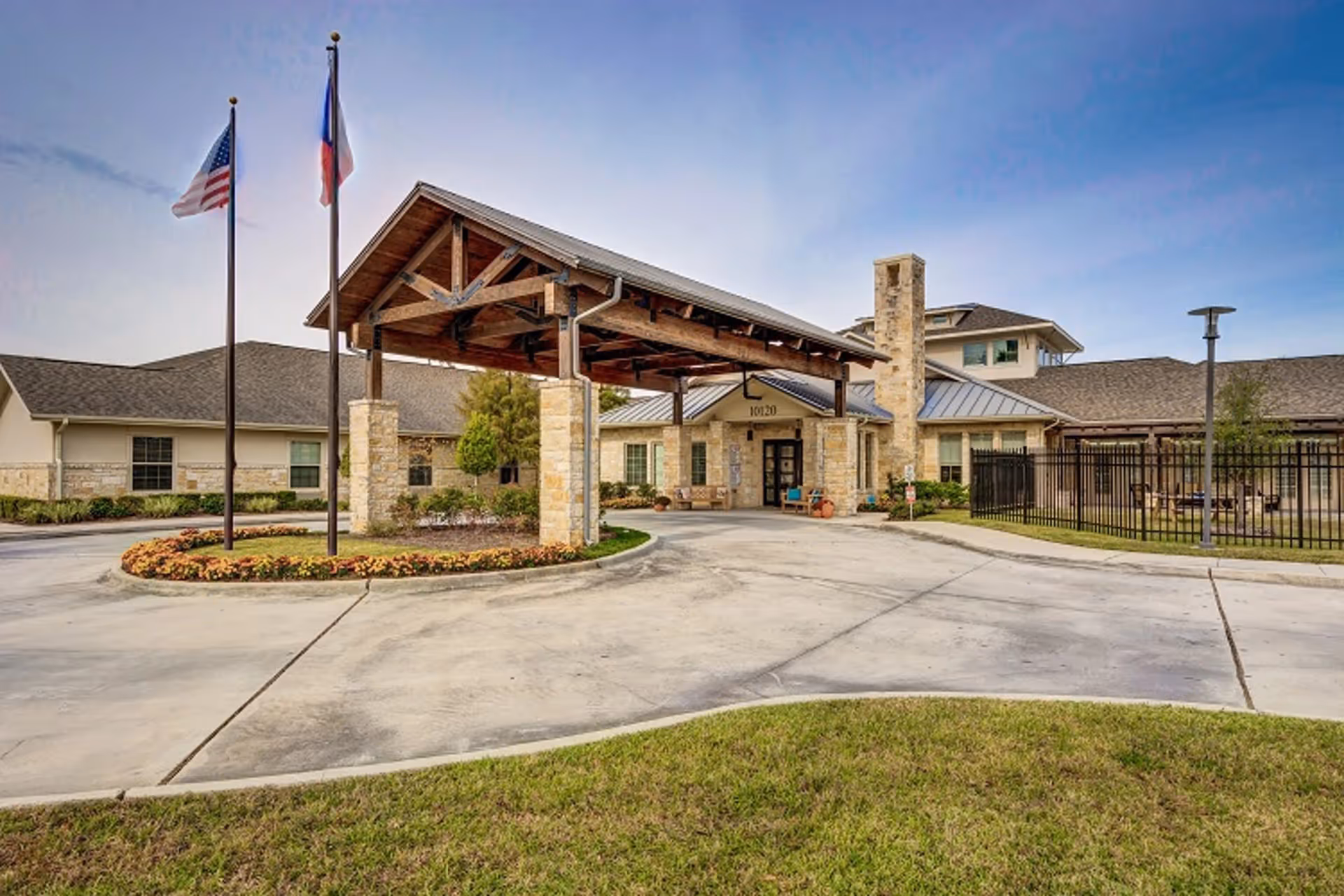 Front entrance of a senior living facility featuring a stone porte-cochere, flagpoles, and a circular driveway.