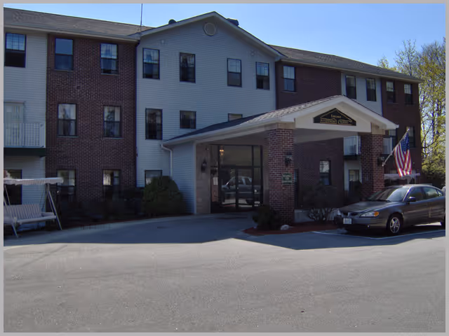 Front entrance of a three-story brick and siding senior living building with a covered porte-cochere, parked car, and an American flag.