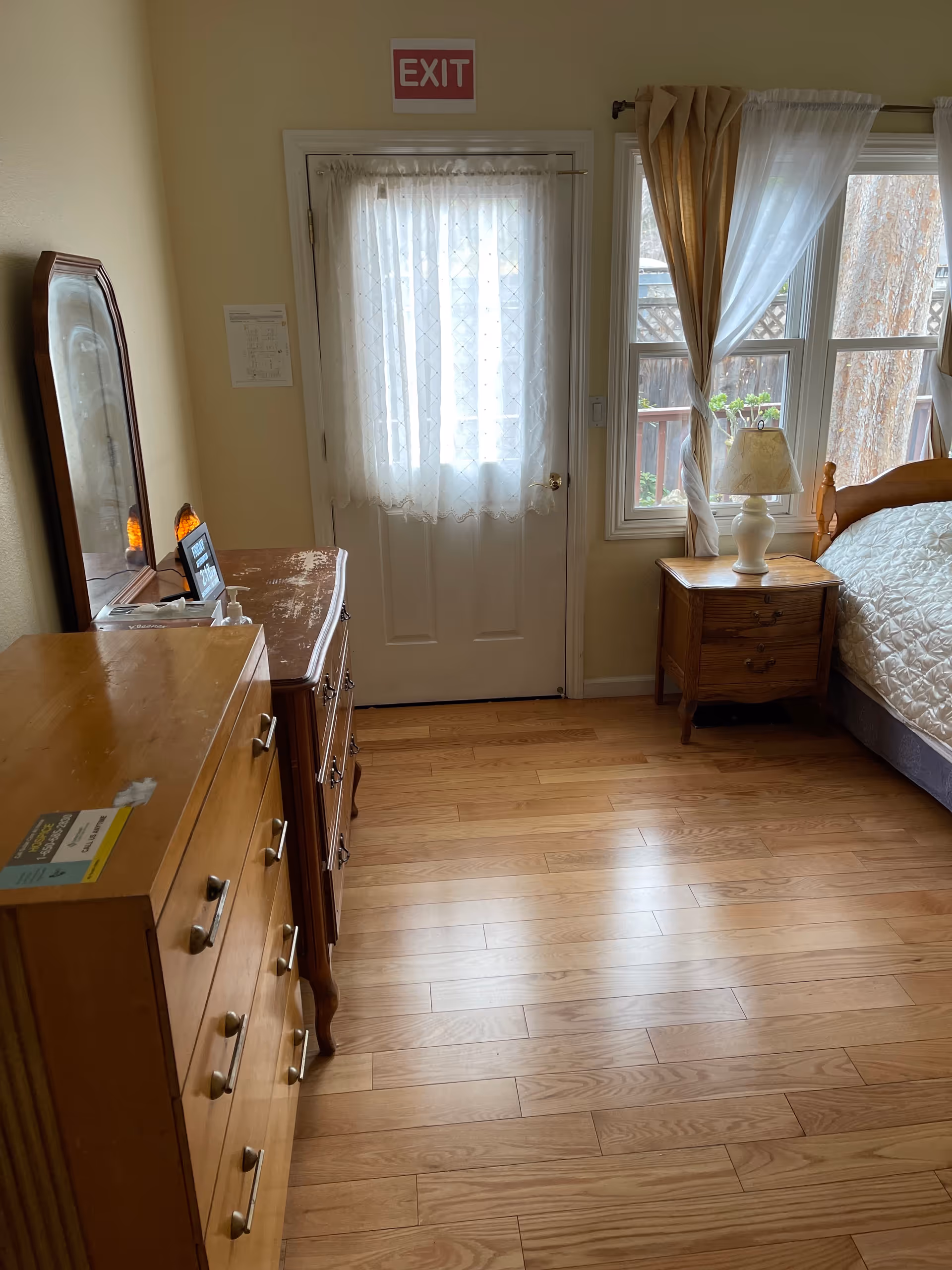 Tidy bedroom interior with dresser, nightstand, bed, window, and exit sign at Orchid Lane facility.