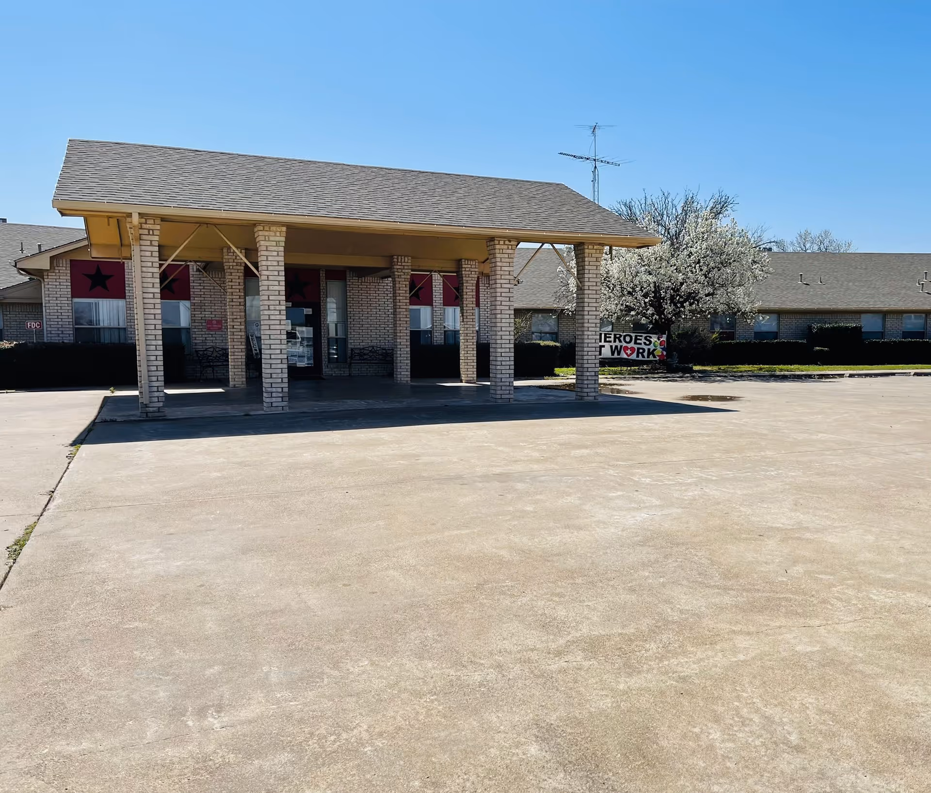 Front exterior view of a single-story brick building with a covered entrance supported by six brick columns. The building has a gray shingled roof and several windows with red decorations featuring black stars. There is a large concrete driveway or parking area in front, and a tree with white blossoms is visible to the right. A sign near the tree reads 'HEROES WORK'.