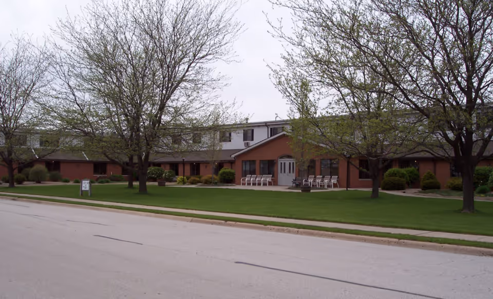 Brick retirement center exterior with central entrance, green lawn, trees, and outdoor chairs.