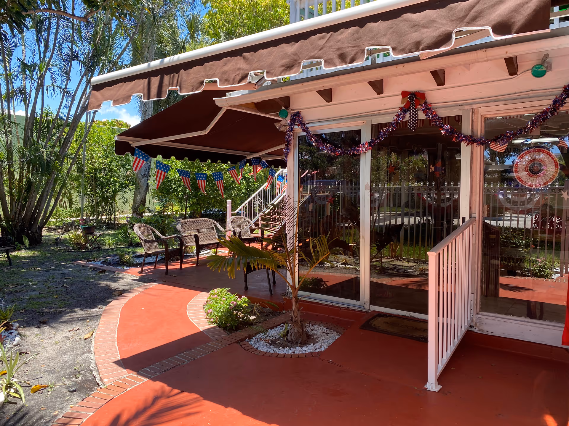 Outdoor patio area with a red concrete floor and brick border, featuring wicker chairs and a small table under a brown awning. The patio is decorated with American flag bunting and garlands. There are palm trees and other greenery surrounding the area, and glass sliding doors lead into the building.