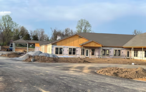 Single-story assisted living building under construction with piles of gravel and a paved driveway in the foreground.