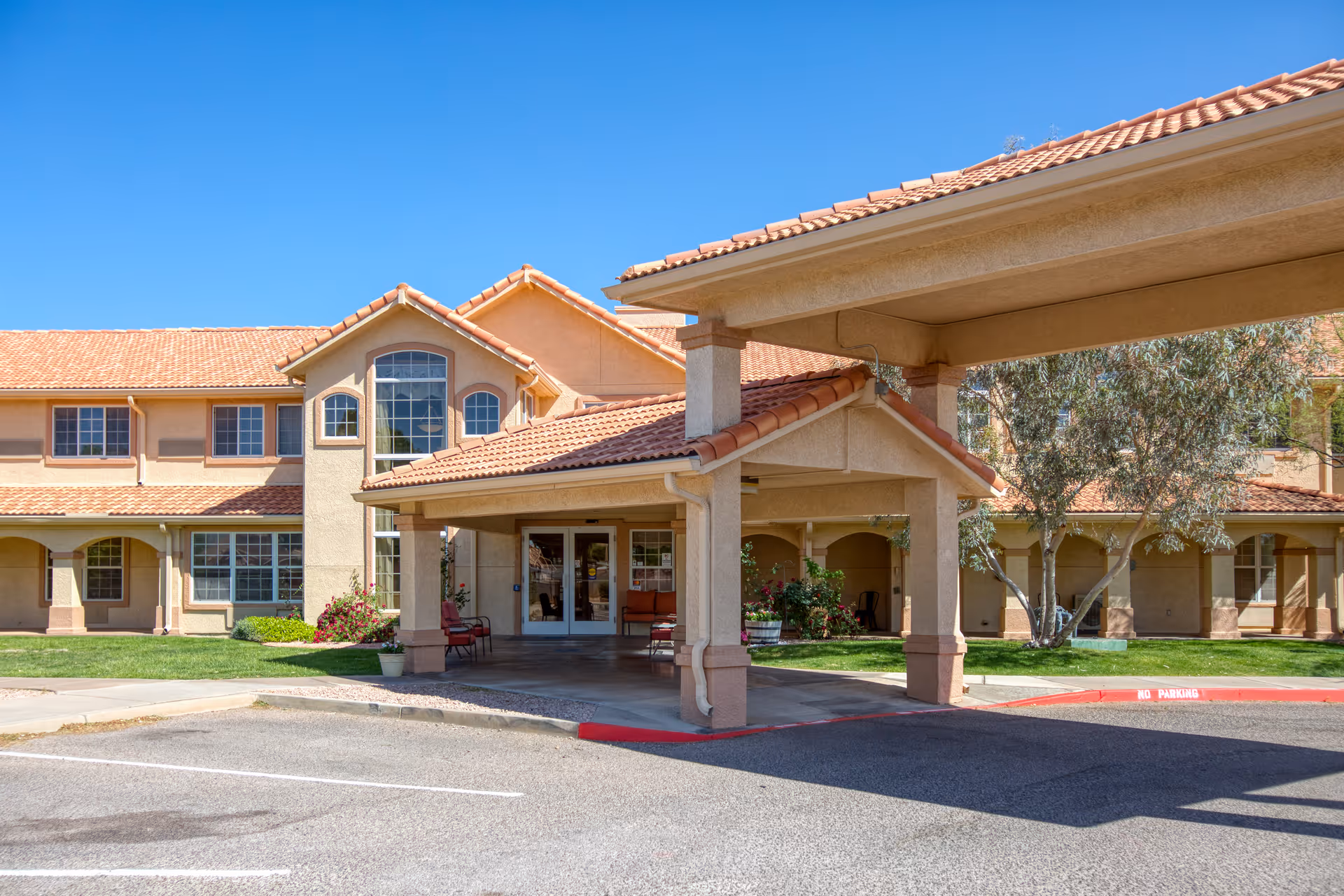 Exterior view of Vista Pointe at Sierra Vista, showing a two-story building with beige walls and a red-tiled roof under a clear blue sky. The entrance features a covered drop-off area with columns and seating, surrounded by green grass and some trees.