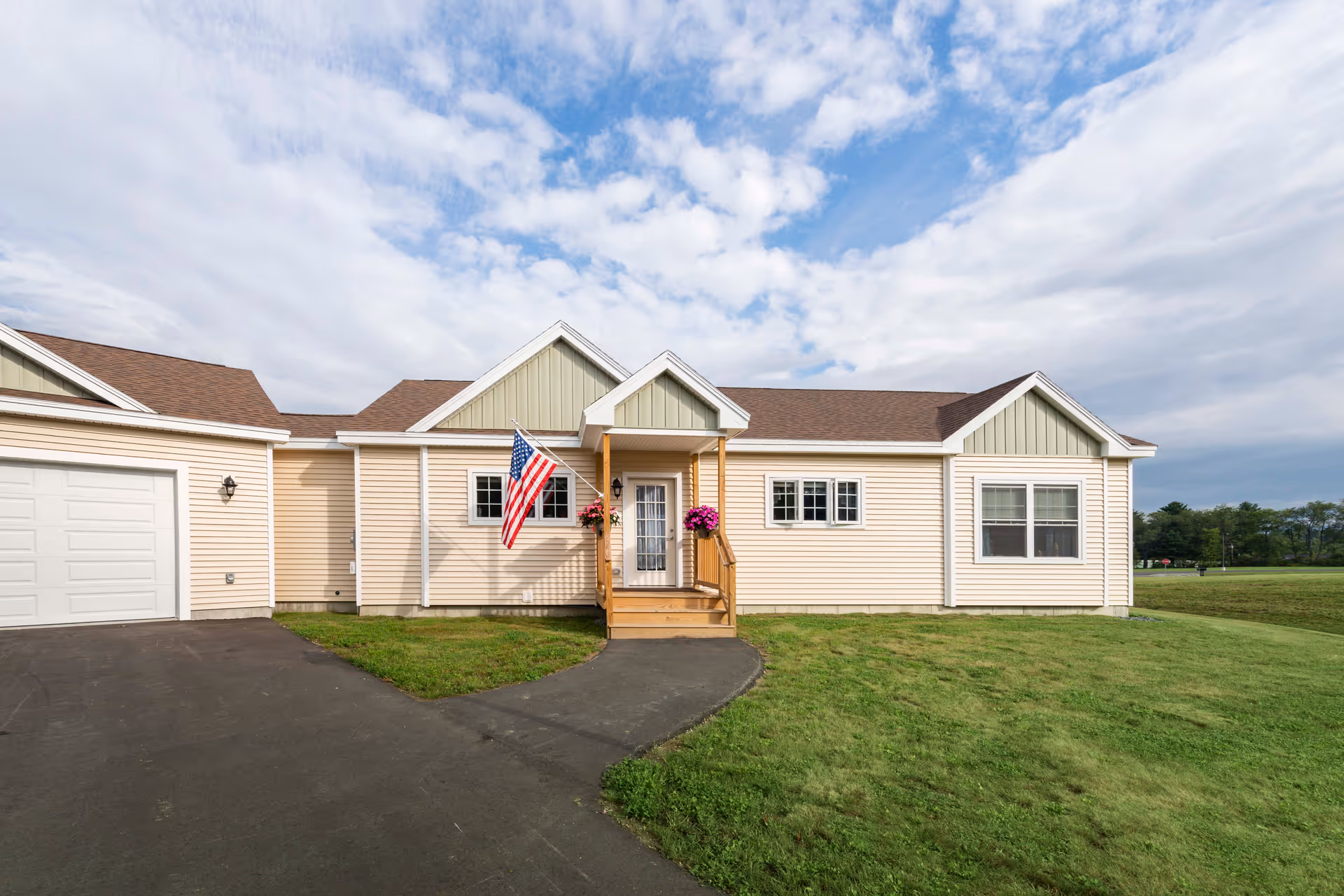 Single-story beige senior living building front with a small porch, American flag, attached garage, and surrounding lawn under a partly cloudy sky.