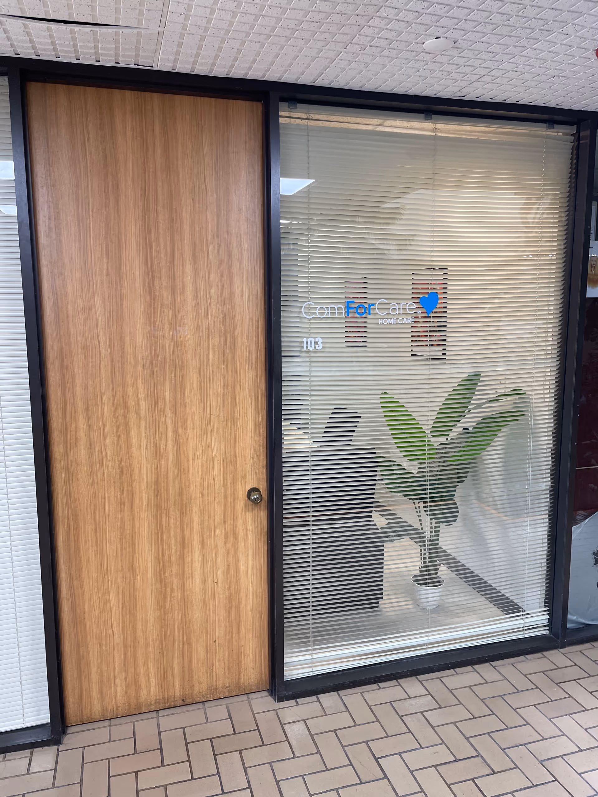 Exterior view of a ComForCare Home Care entrance with wood door, blinds, and plant visible.