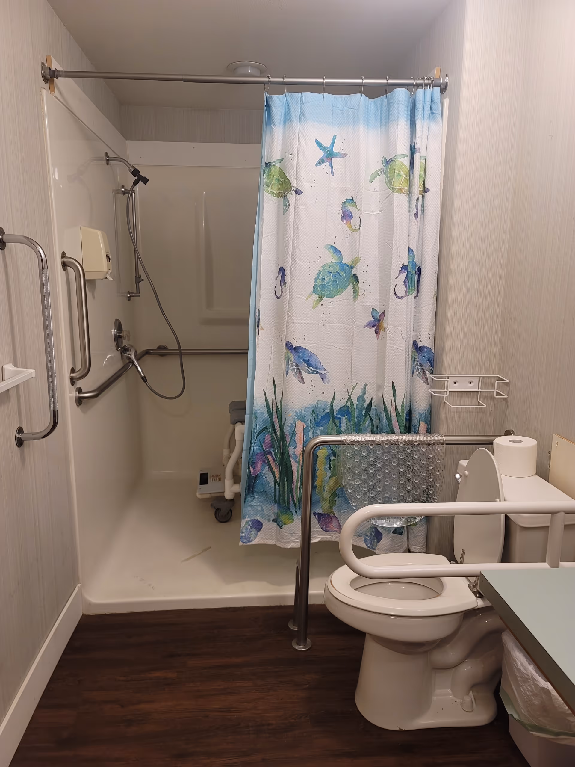 Accessible bathroom in a senior facility with shower, grab bars, toilet, and sea-themed curtain.