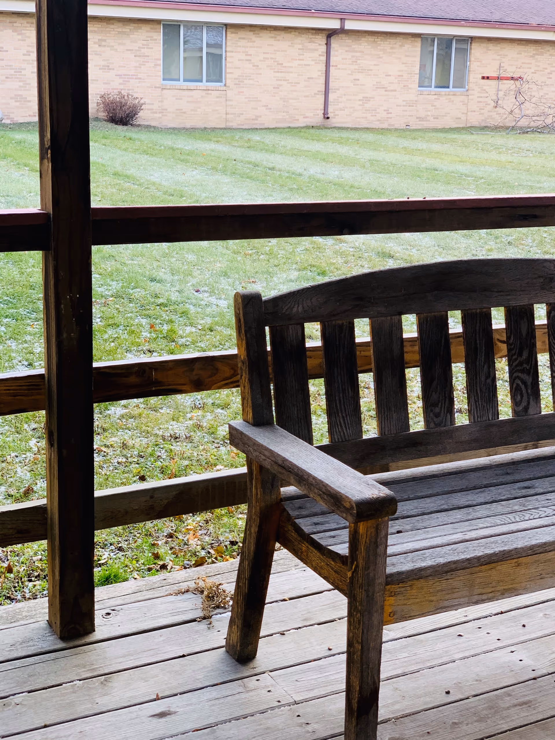 Wooden bench on a covered porch overlooking a grassy courtyard and a brick building with windows.
