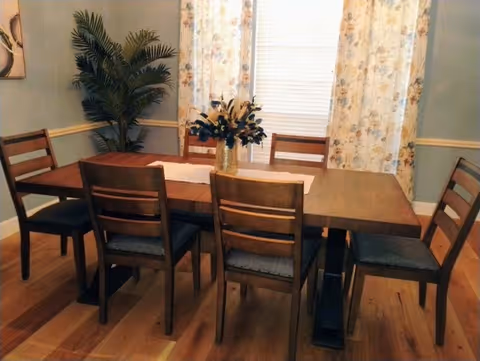 A dining room with a wooden table and six matching wooden chairs with dark cushions. A vase with flowers is placed in the center of the table. Behind the table, there is a window with floral curtains and a white blind. A large green potted plant is positioned in the corner of the room.