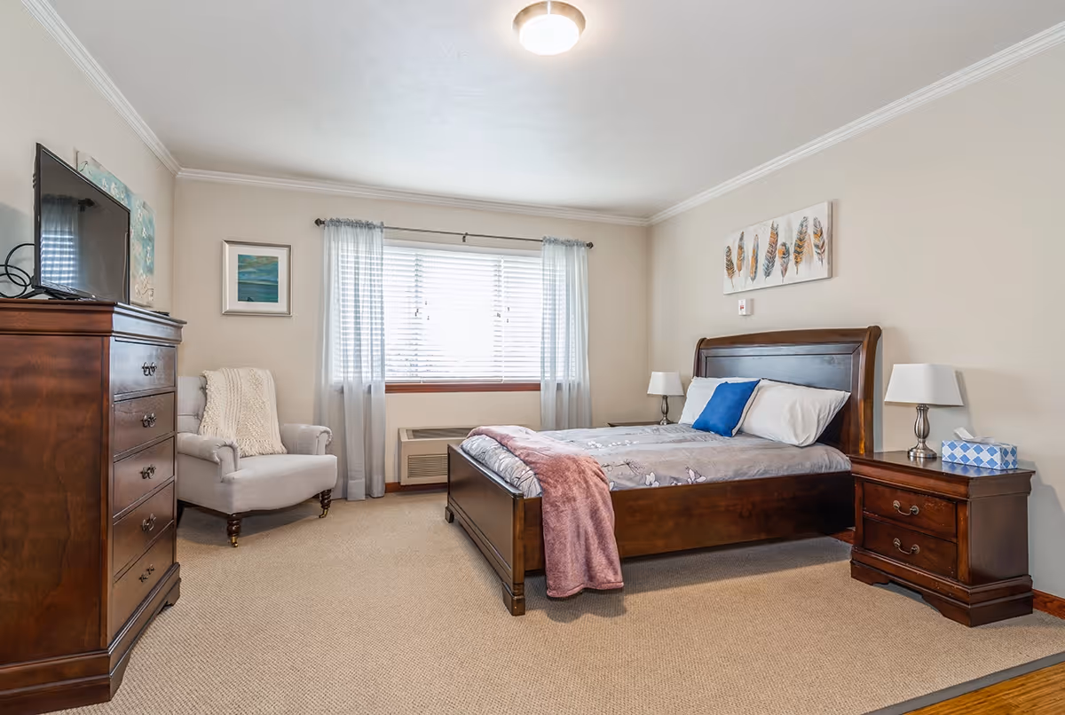 Spacious bedroom with a wooden bed, matching nightstands and dresser, an upholstered armchair, and a window with curtains.