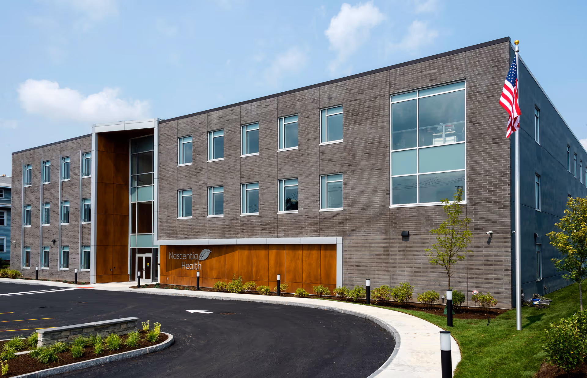 Modern brick Nascentia Health facility front with glass entrance, curved driveway, and American flag.