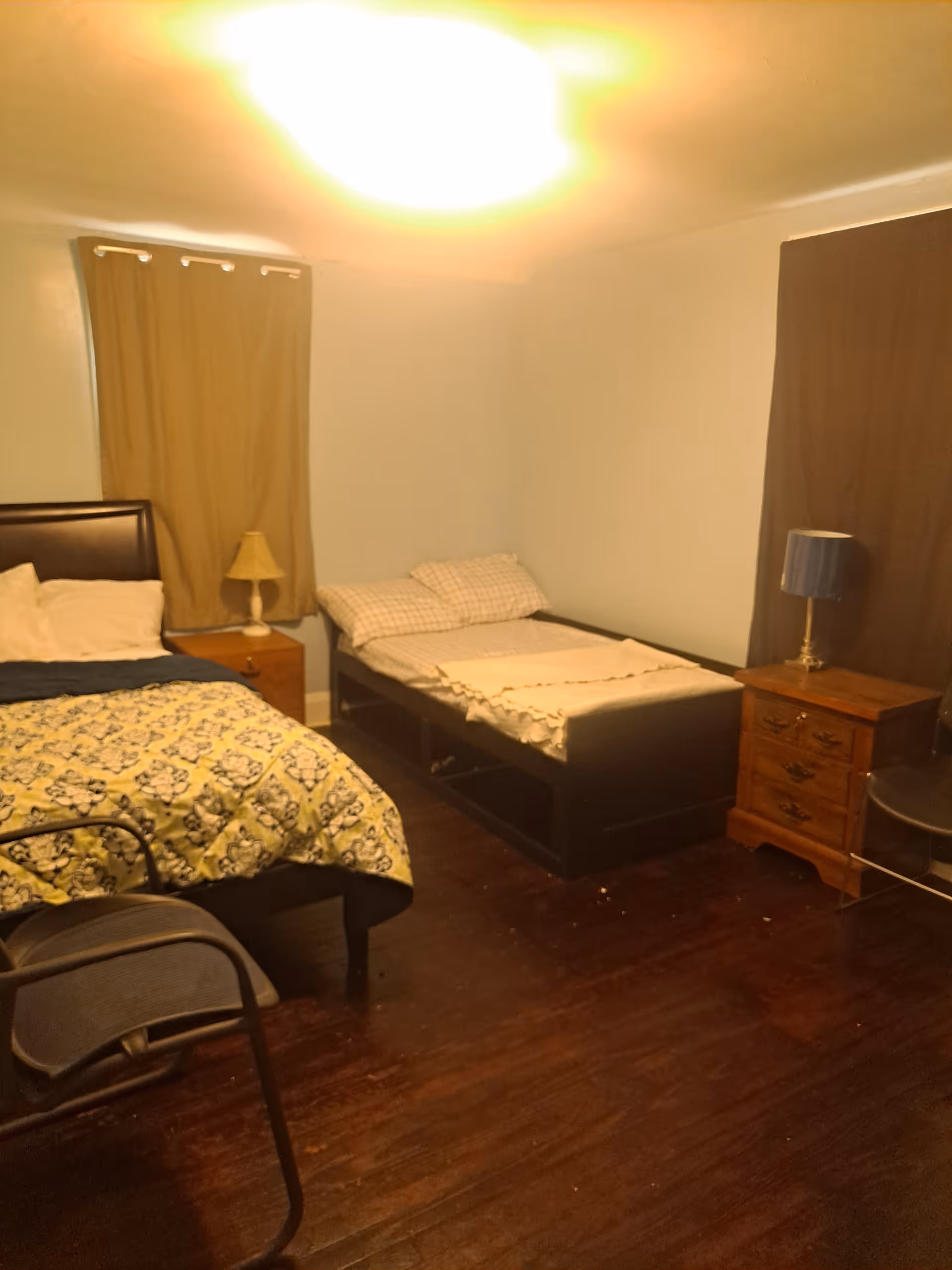 Two beds and wooden furniture fill a small, dimly lit bedroom.