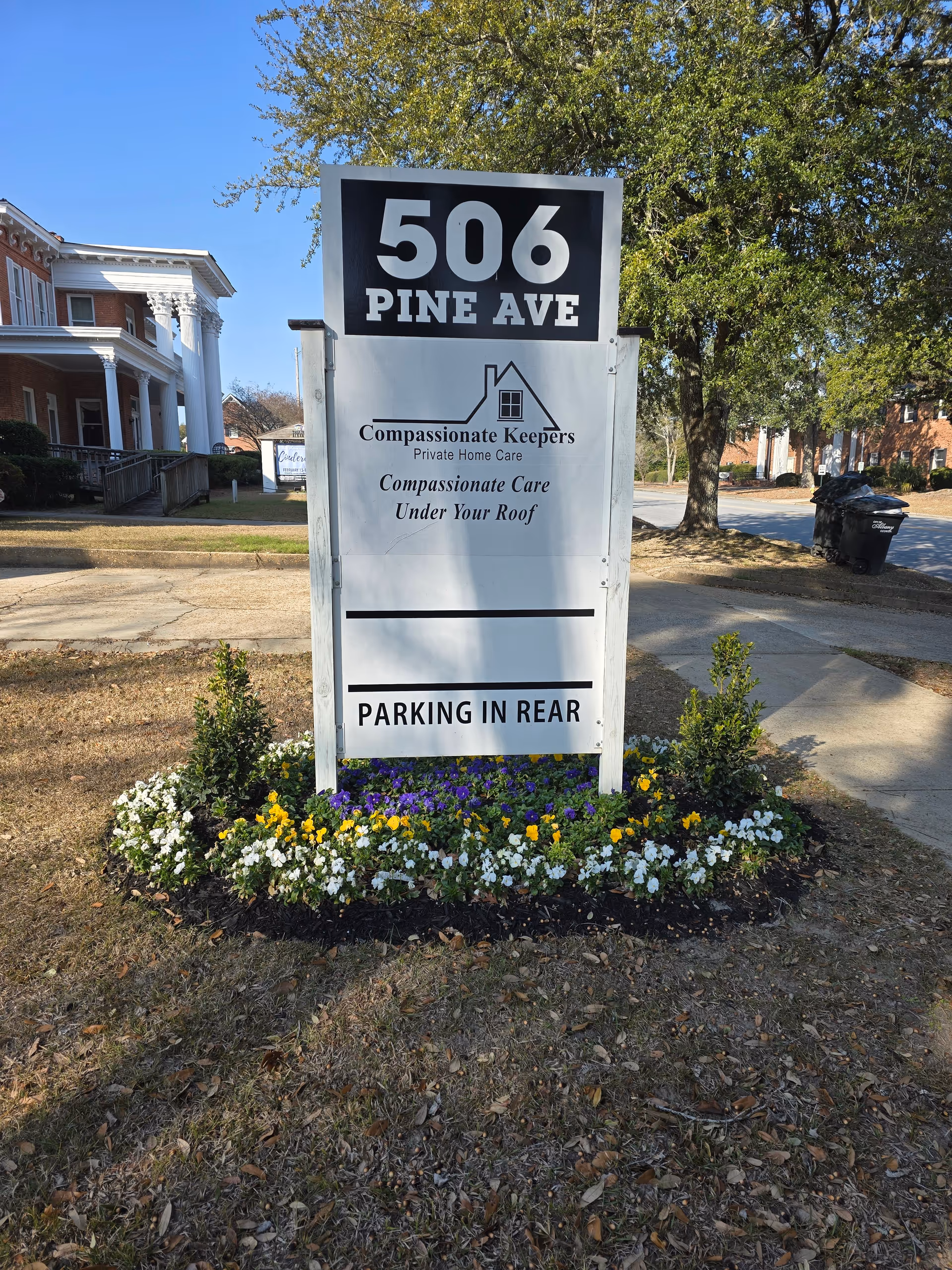 Tall exterior sign for Compassionate Keepers Home Care at 506 Pine Ave surrounded by flowers.