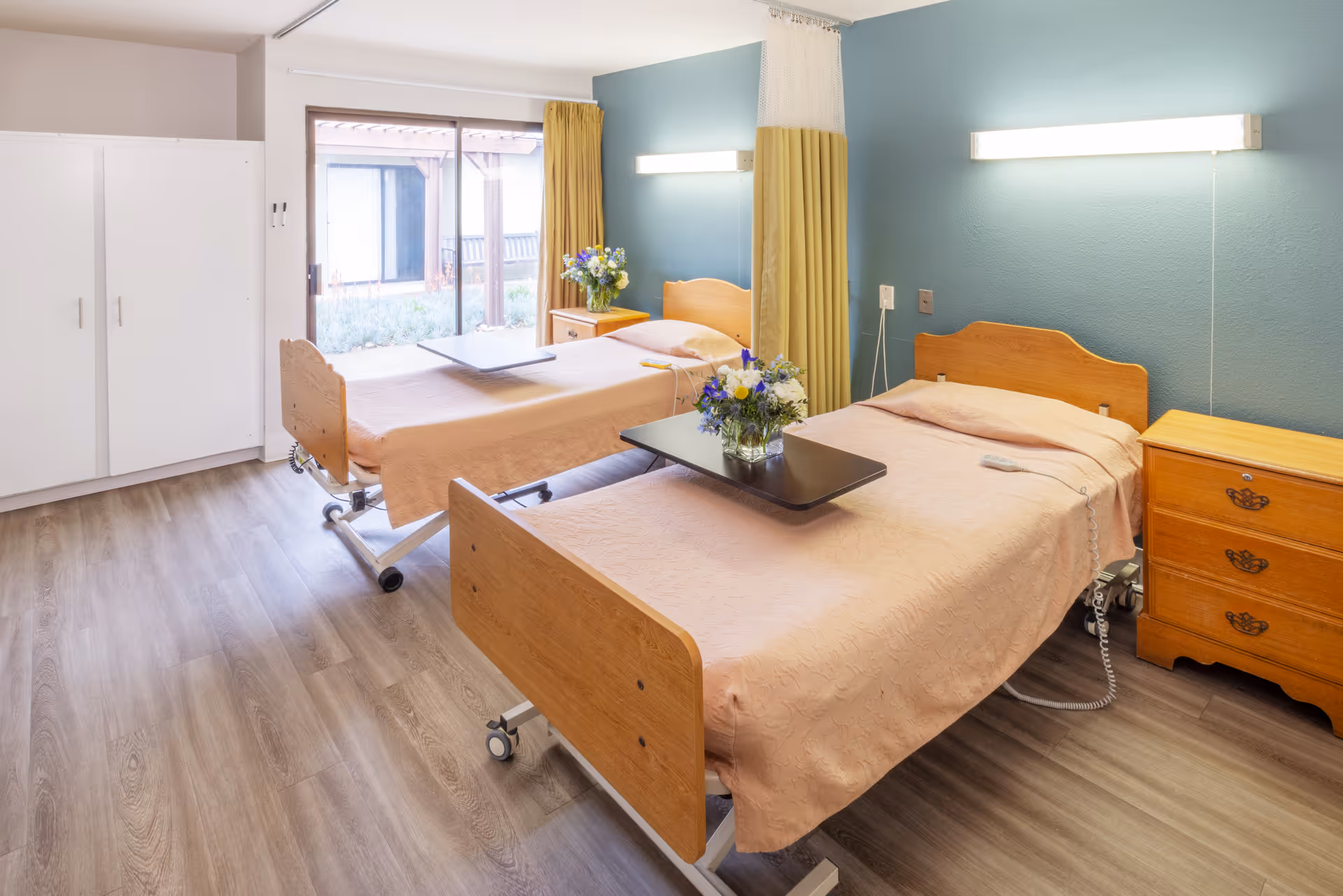 A bright and clean bedroom in a healthcare facility with two hospital-style beds covered in light peach bedspreads. Each bed has a wooden headboard and footboard, a bedside table with drawers, and a small table with a vase of flowers. The room has wood-look flooring, a large window with yellow curtains, and a blue accent wall with mounted lights above each bed.
