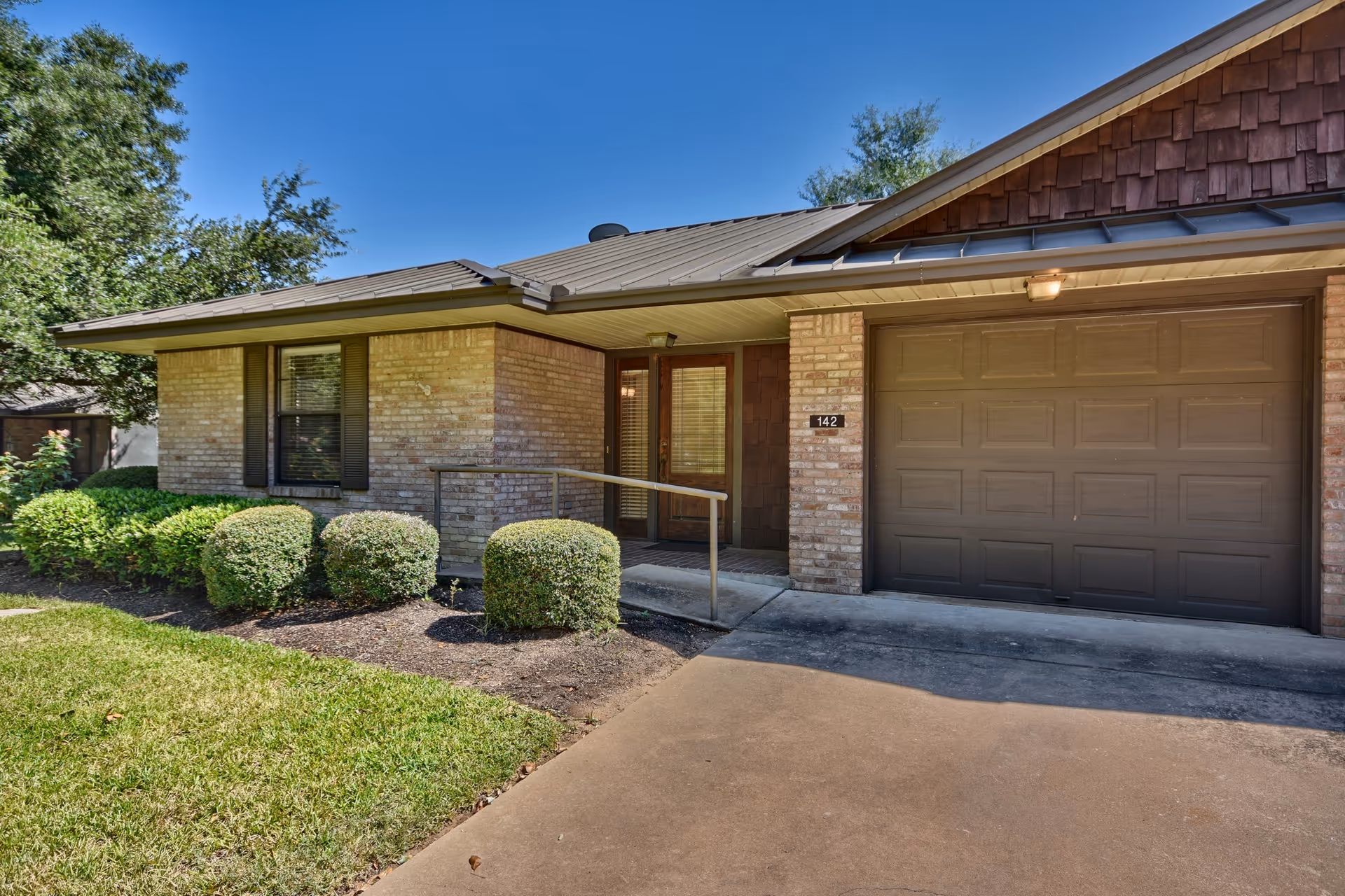 Exterior view of a single-story brick house with a brown garage door, a small front porch with a metal handrail, neatly trimmed bushes, and a green lawn under a clear blue sky.