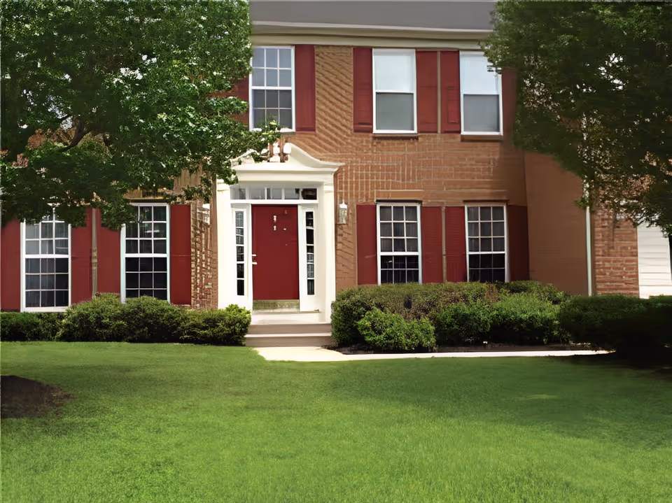 Two-story brick house front with a red door, red shutters, and a green lawn.