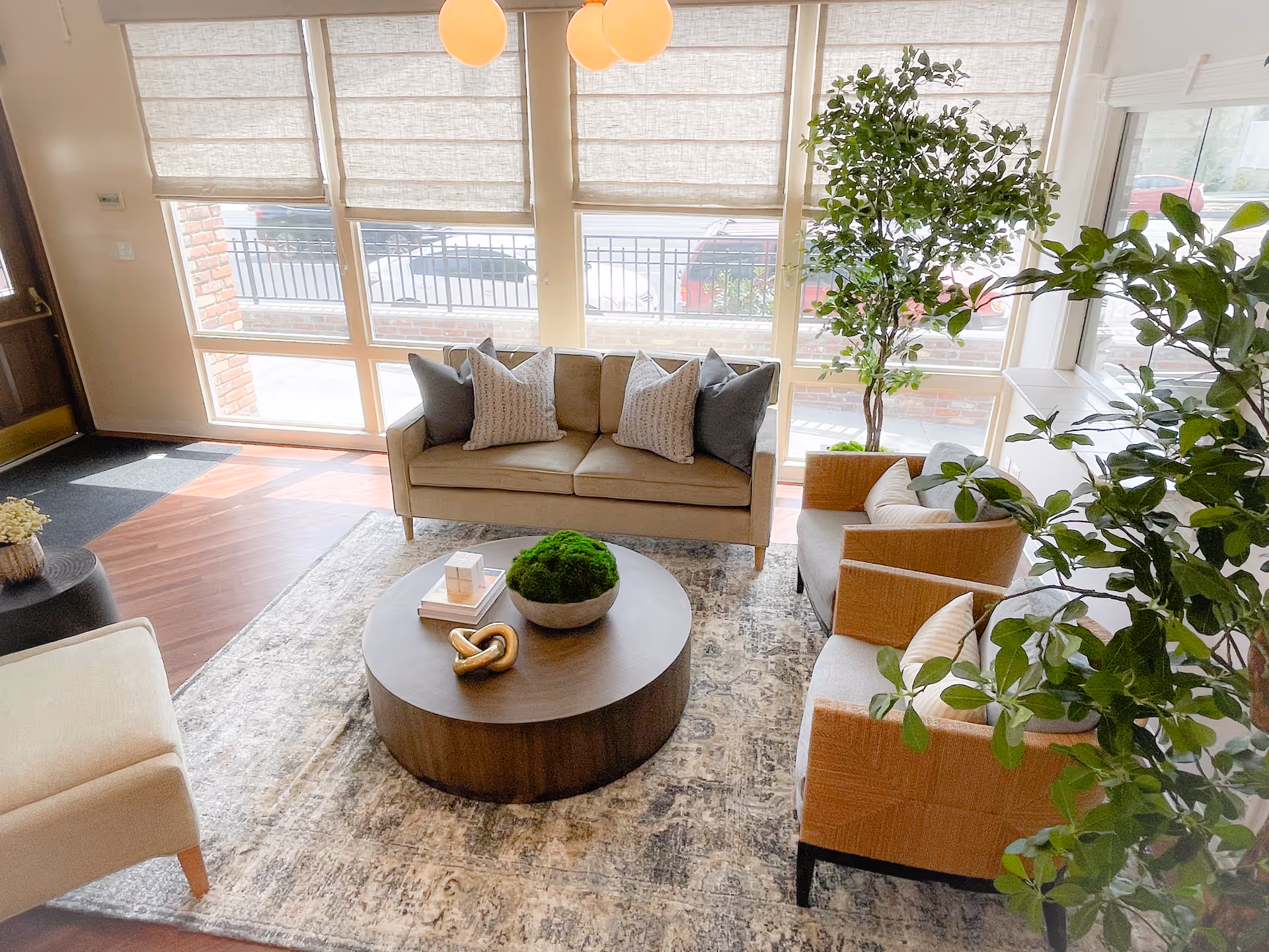 A bright and cozy living room area with a beige sofa adorned with four pillows, two armchairs with cushions, a round wooden coffee table with decorative items, a patterned rug, large windows with blinds letting in natural light, and several green plants adding a touch of nature.