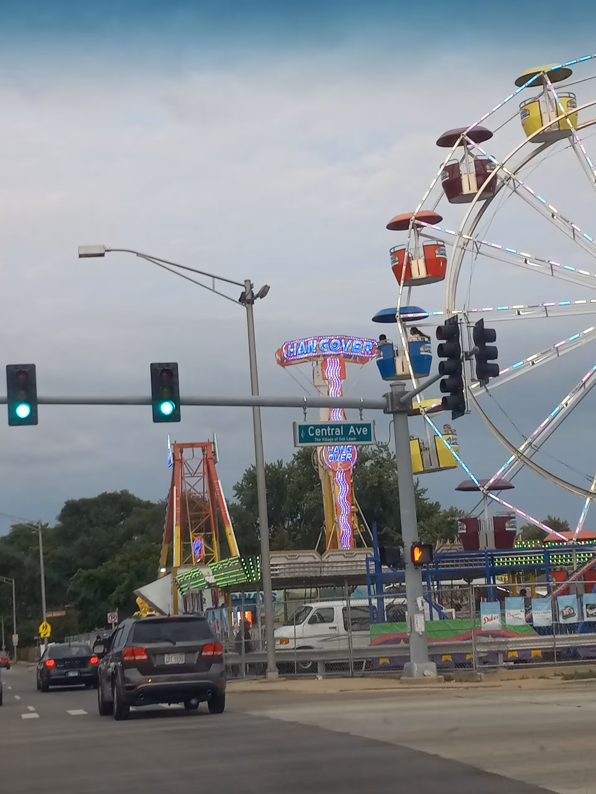 Street scene with a large Ferris wheel and carnival rides beside traffic signals.