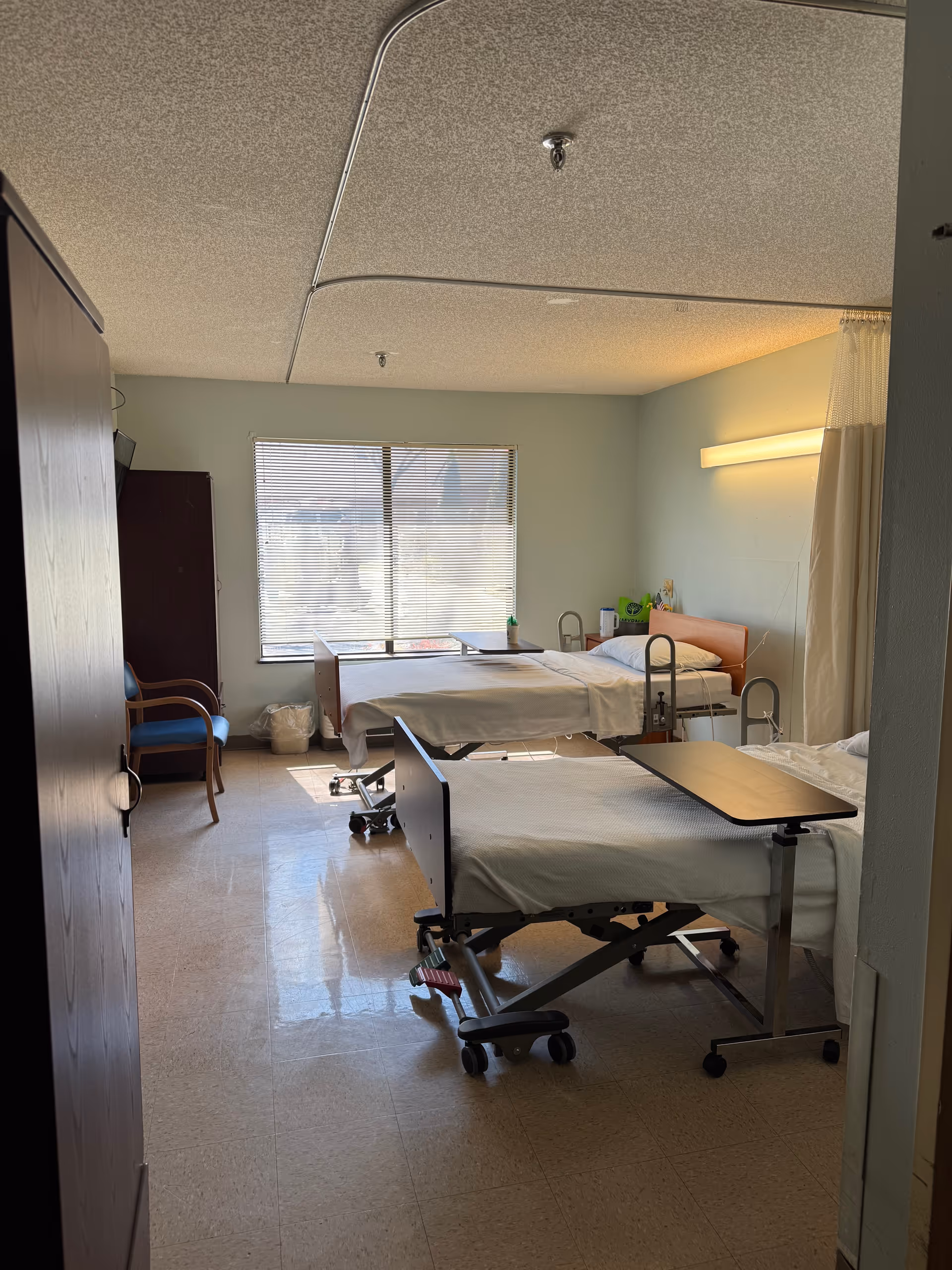 Bright patient room with two beds, window, and medical furniture.