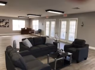 A spacious living room area with three dark gray sofas arranged around a glass coffee table. The room has light-colored wood flooring, large windows, and glass doors letting in natural light. There are a few chairs and a small table in the background near the windows, and neutral-colored walls with minimal decor.