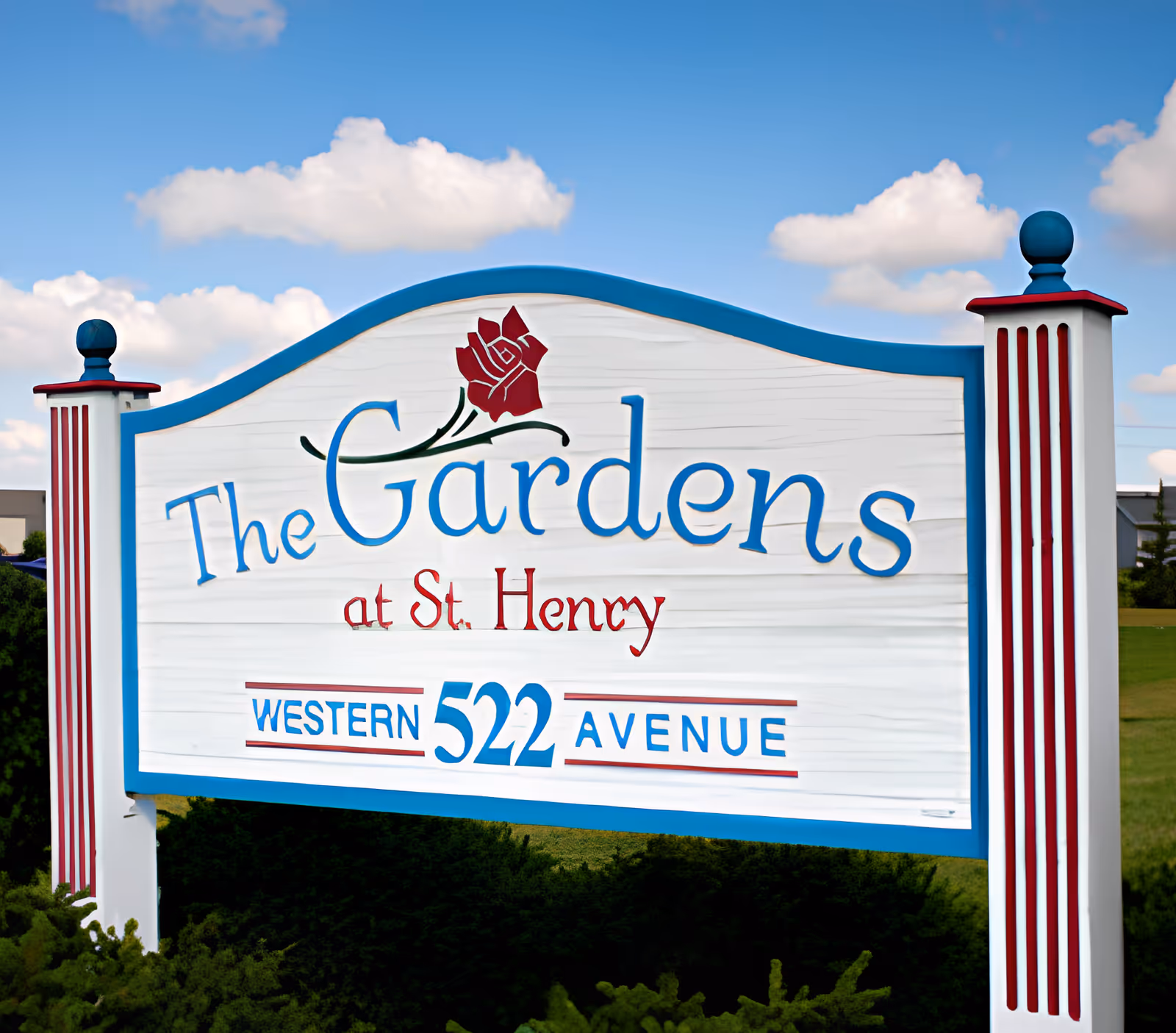White and blue entrance sign that reads "The Gardens at St. Henry" with the address "Western 522 Avenue" against a blue sky.