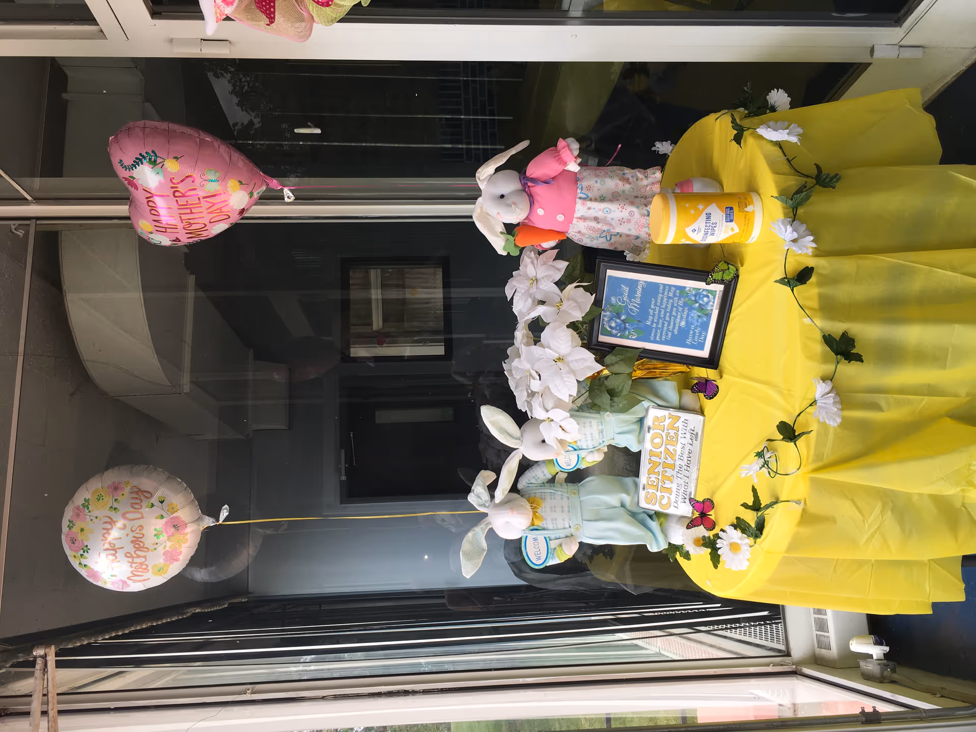 Entrance display with yellow tablecloth, bunny decor, and balloons outside Passion and Caring Home.