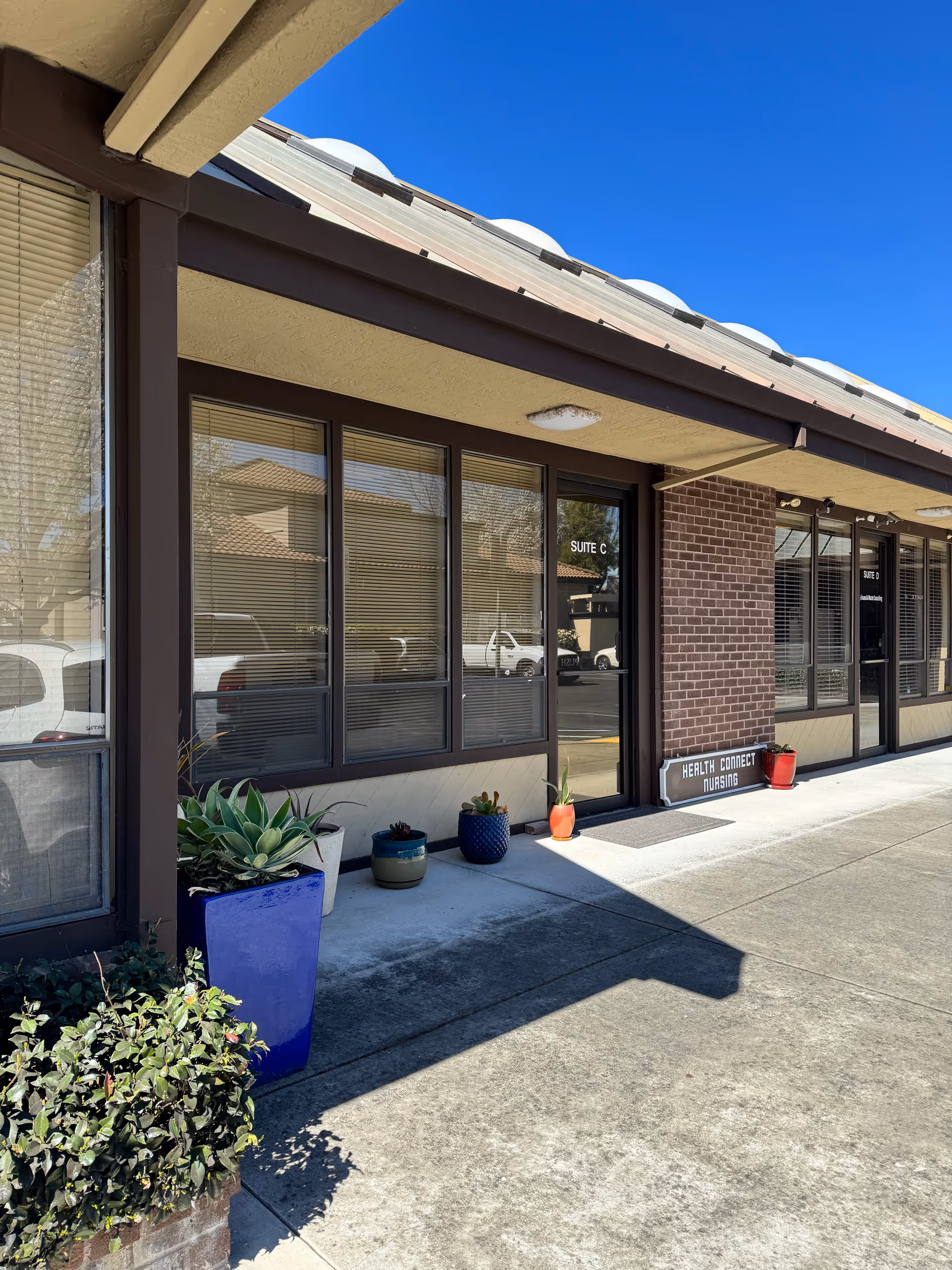 Exterior storefront of Health Connect Nursing, brick columns, large windows, and potted plants along the sidewalk.