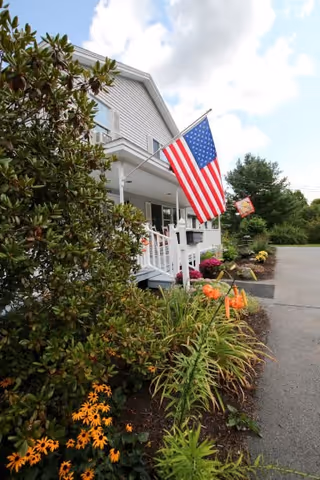 Front porch of a white retirement home with an American flag, landscaped flower beds, and a driveway.