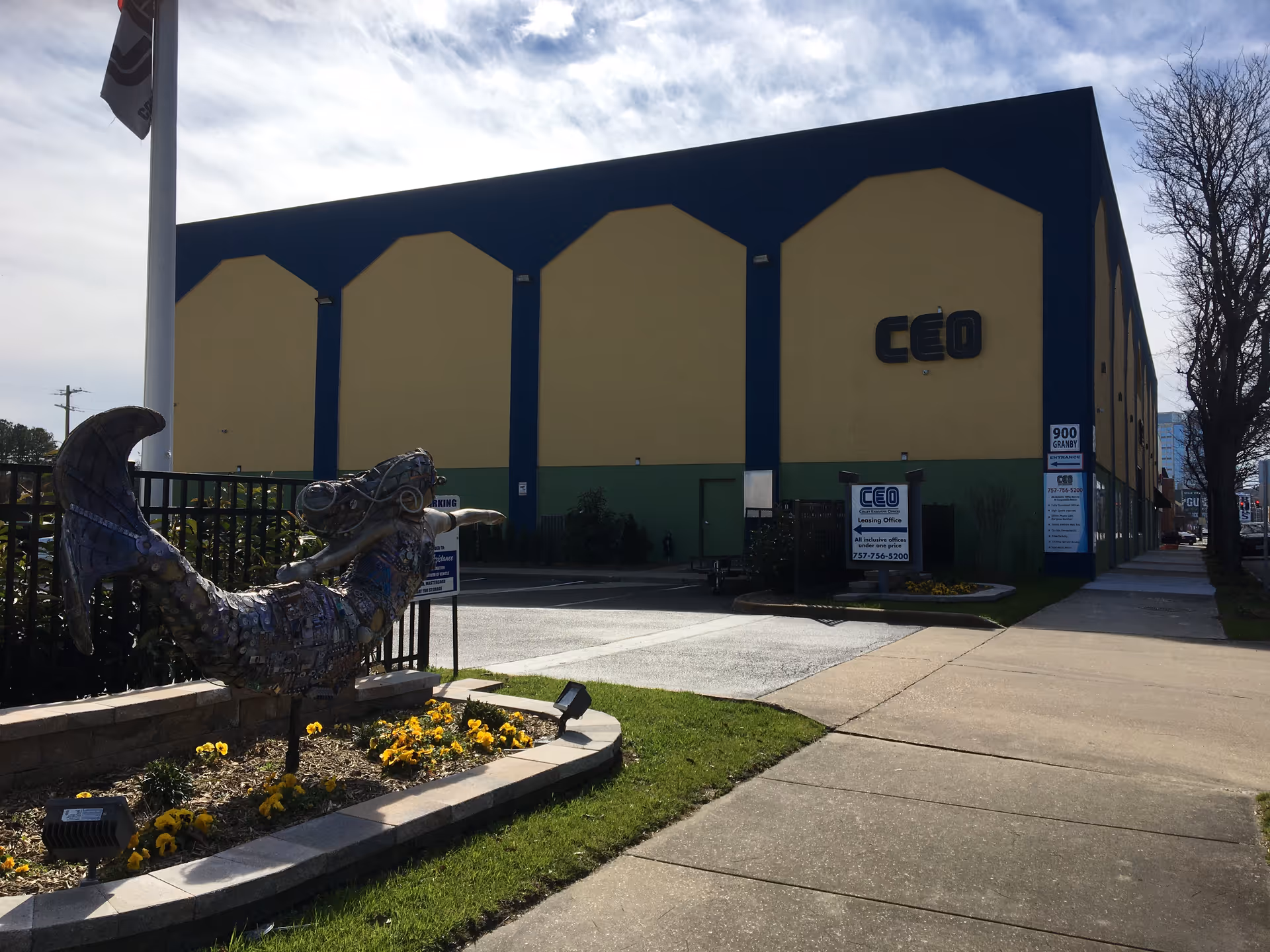 Exterior view of a large blue and yellow building with a metal sculpture and flowers.