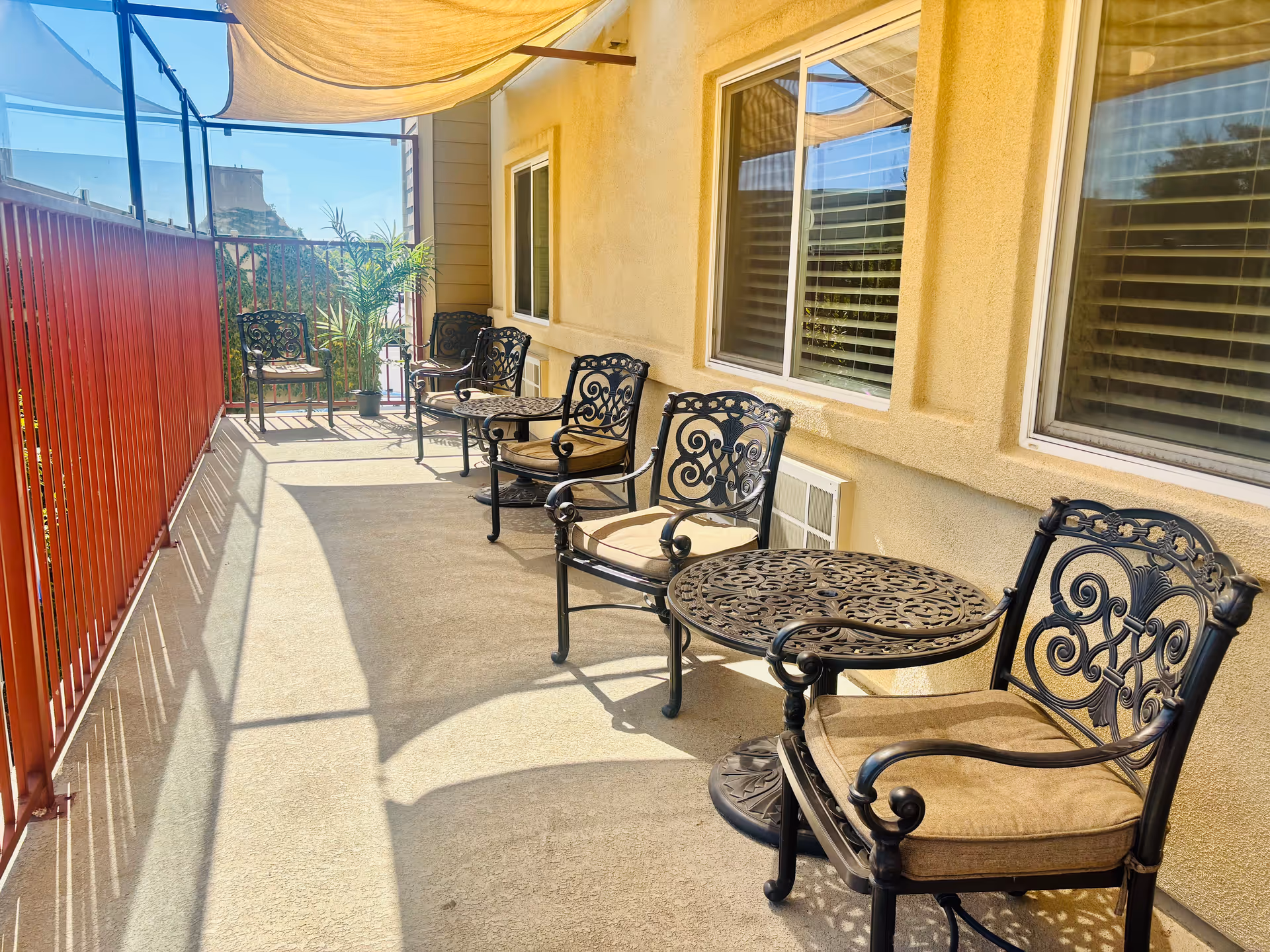 Outdoor patio with ornate metal chairs and tables along a beige building, shaded by a canopy.